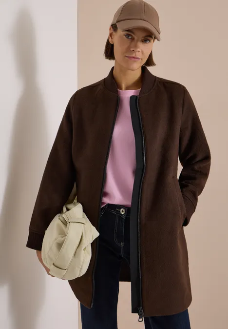 Femme en manteau marron, haut rose, pantalon foncé, sac beige et casquette.