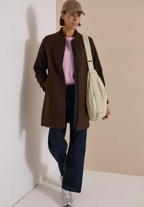 Femme en manteau marron, pull rose, jean large, casquette et grand sac, posant devant un mur neutre.