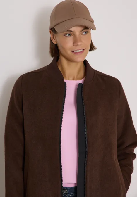 Femme portant une veste marron, t-shirt rose et casquette, souriante.