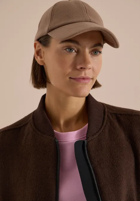 Femme portant une casquette brune, une veste marron et un t-shirt rose, regard latéral.