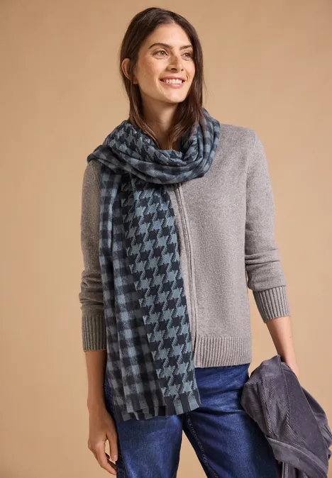 Przytulny szalik z frędzlami i wzorami dark navy blue Kobieta w szarym swetrze z niebiesko-czarnym szalem pepitkowym, trzyma kurtkę i uśmiecha się.