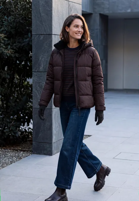 Femme en doudoune marron et jeans marchant en souriant devant un bâtiment moderne.