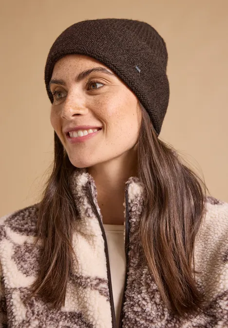 Eine lächelnde Frau mit braunen Haaren, trägt eine dunkelbraune Strickmütze und eine gemusterte Fleecejacke.
