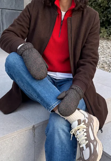 Moufles doublées à motif léopard Black Person sitzt draußen auf einer Steinbank, trägt braunen Mantel, rotes Oberteil, graue Fäustlinge, Jeans und weiße Sneaker.