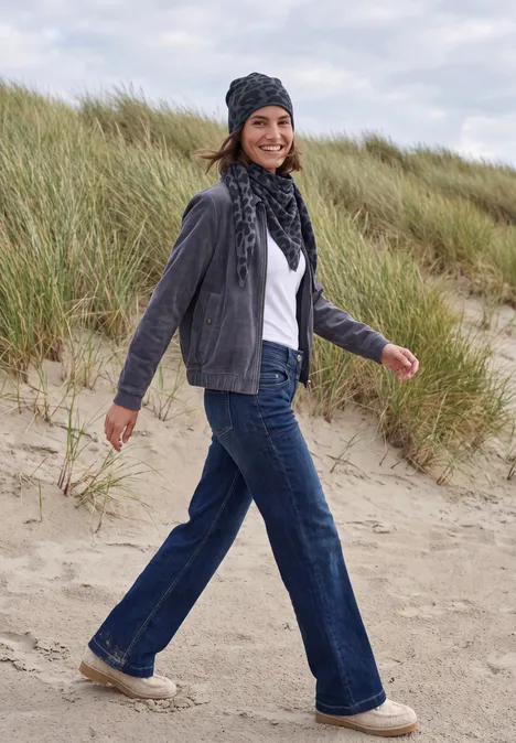 Eine Frau spaziert am Strand vor Dünen, trägt Jeans, graue Jacke, Schal und Mütze.