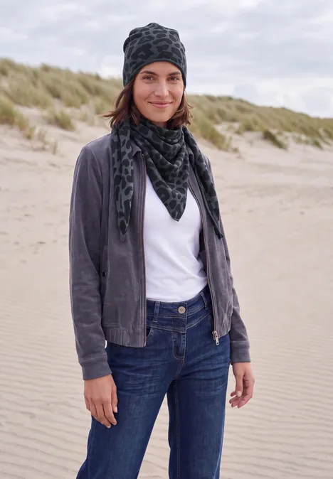 Eine Frau steht an einem Sandstrand mit Dünen und trägt Mütze, Schal, graue Jacke, weißes T-Shirt und Jeans.