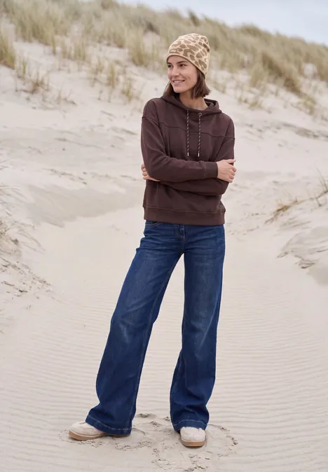 Junge Frau im Hoodie und Strickmütze steht in Dünen am Strand.
