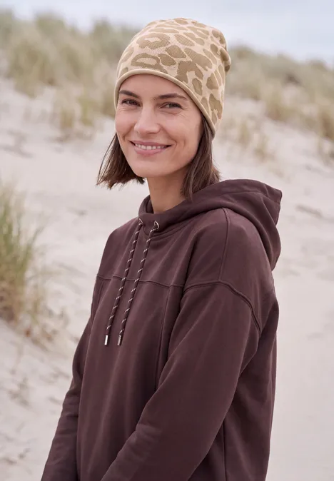Eine lächelnde Frau in braunem Hoodie und beige-gemustertem Mütze an einem Dünenstrand.