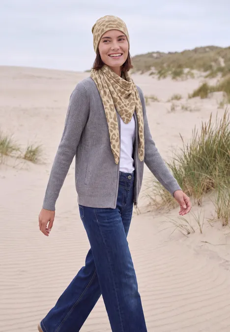 Driehoekige sjaal met luipaardprint powder beige Een glimlachende vrouw wandelt over het duinstrand, draagt spijkerbroek, grijze cardigan en bijpassende muts met sjaal.
