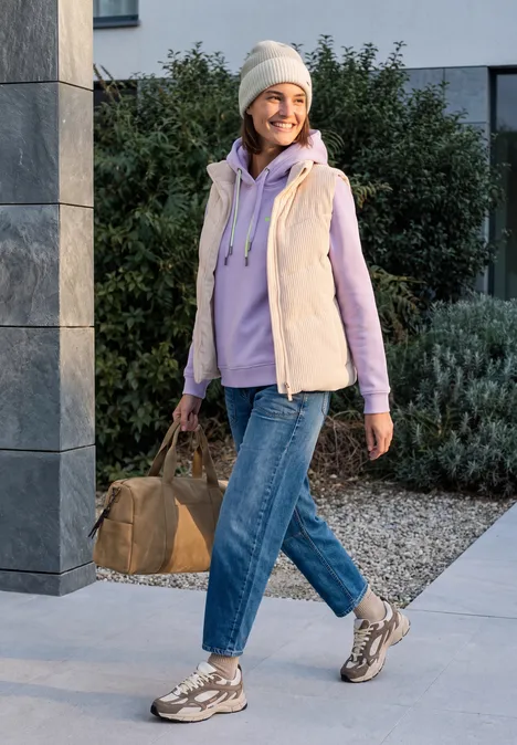 Beanie met ribbelsteek in tweekleurige uitvoering powder beige melange Vrolijke vrouw met muts, bodywarmer en jeans wandelt buiten met tas.