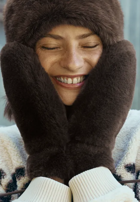 Une femme souriante, emmitouflée dans un bonnet et des moufles en fourrure, se couvre le visage.