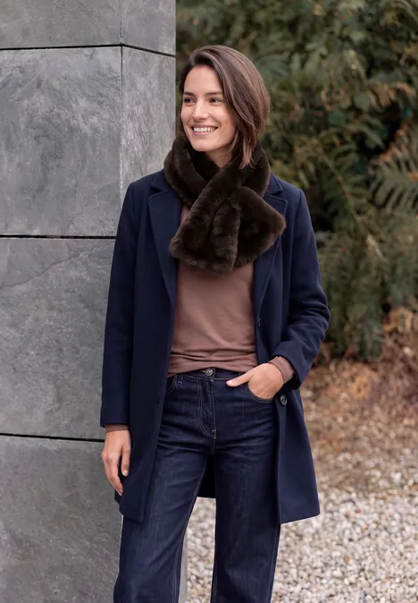 Eine lächelnde Frau in Jeans, braunem Pullover, blauem Mantel und braunem Pelzschal steht draußen vor einer Wand.