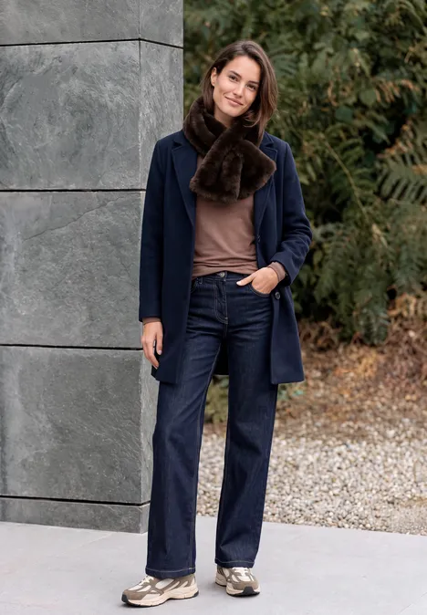 Eine Frau in Mantel, Schal und Jeans steht vor einer Mauer und Grün, lächelt locker.