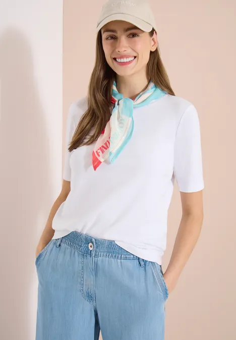 Een vrouw draagt een wit T-shirt, een kleurrijke zijden sjaal en een lichtblauwe, hoog getailleerde broek.