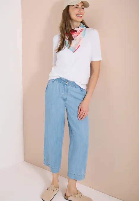 Vrouw met wit t-shirt, kleurrijke sjaal, wijde lichtblauwe jeans-culotte en sandalen tegen lichte achtergrond.