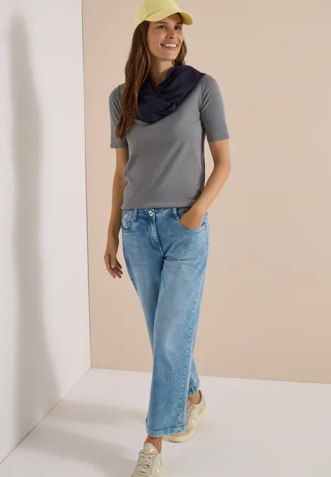 Frau in grauem T-Shirt, blauen Jeans, gelber Kappe und Schal vor neutralem Hintergrund.