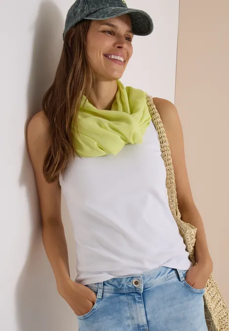 Écharpe Loop unie citrin green Femme souriante en casquette, débardeur blanc, foulard vert, jean et sac en paille adossée à un mur.