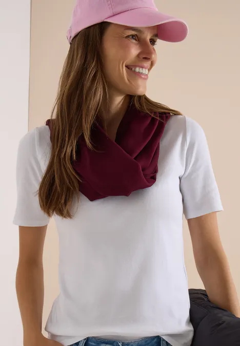 Écharpe Loop unie red wine Une femme souriante en t-shirt blanc, casquette rose et foulard bordeaux posé sur les épaules.