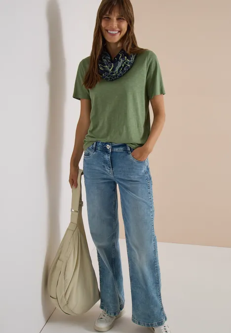 Écharpe Loop à motif léopard dried olive green Femme en t-shirt vert, jean évasé, foulard au cou et grand sac beige sur fond neutre.