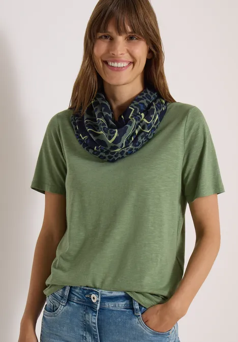 Écharpe Loop à motif léopard dried olive green Une femme souriante porte un t-shirt vert et un foulard tubulaire à motifs sur fond clair.