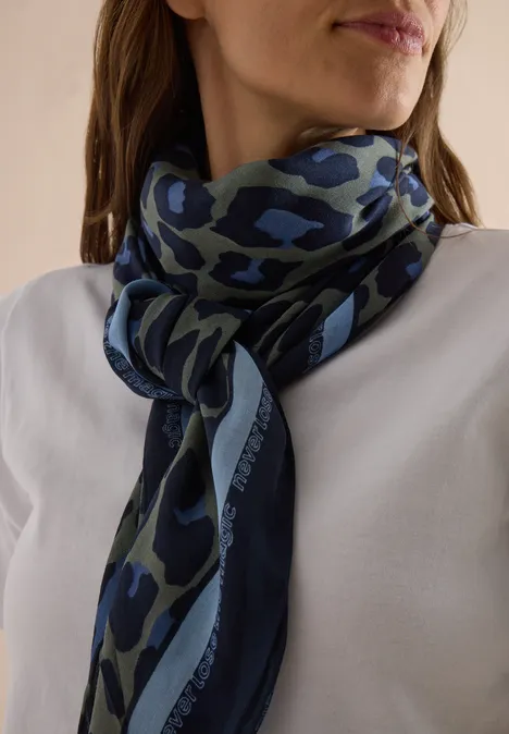 Vrouw in wit shirt met een blauw-groene camouflage-sjaal los om de hals geknoopt.