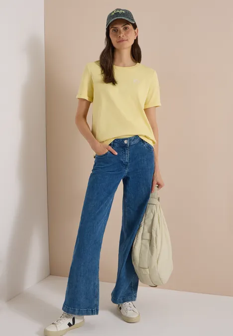 Pet met leeuwenpatroon en slogan dried olive green Frau mit gelbem T-Shirt, weiten Jeans, weißer Tasche und grüner Baseballkappe vor heller Wand.