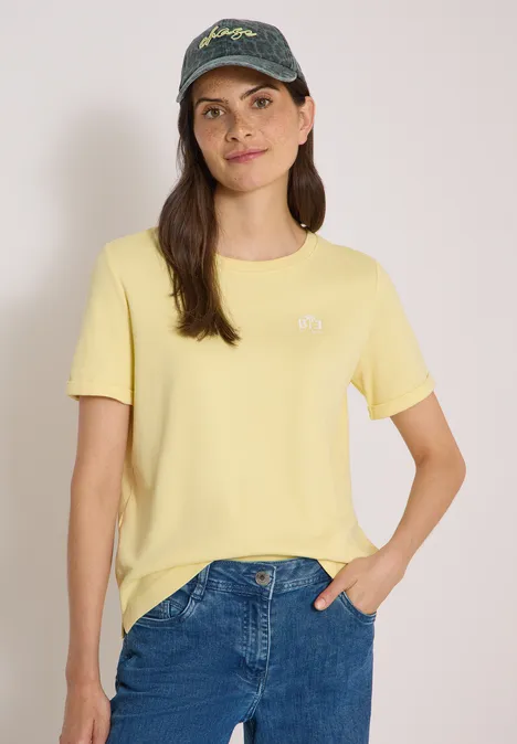 Pet met leeuwenpatroon en slogan dried olive green Eine Frau mit gelbem T-Shirt, blauer Jeans und Baseballcap lächelt vor neutralem Hintergrund.