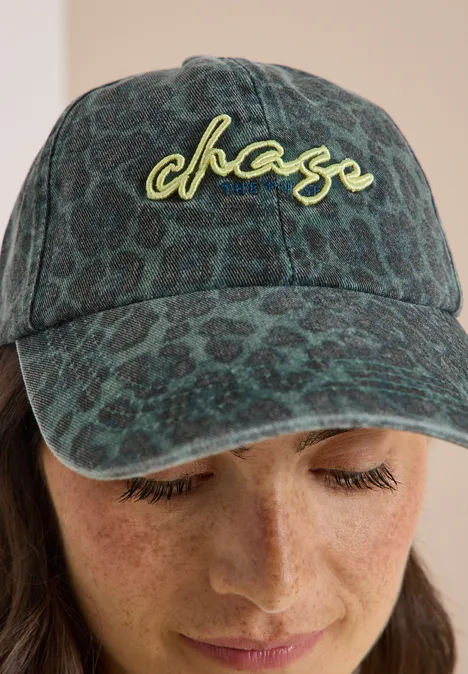 Pet met leeuwenpatroon en slogan dried olive green Person mit grün gemustertem Cap mit chase-Logo und Sommersprossen blickt nach unten.