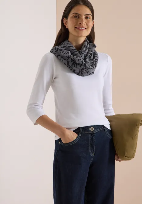 Femme souriante en tee-shirt blanc à manches longues, jean foncé et écharpe grise, tenant un manteau.