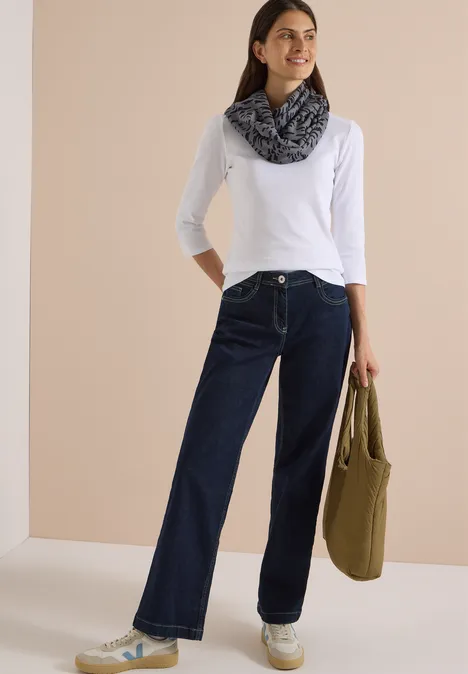 Femme en t-shirt blanc à manches longues, jean ample foncé, foulard à motifs et sac kaki sur fond neutre.