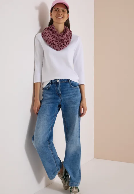 Femme en jean, haut blanc, casquette et écharpe roses, souriante, adossée à un mur clair.