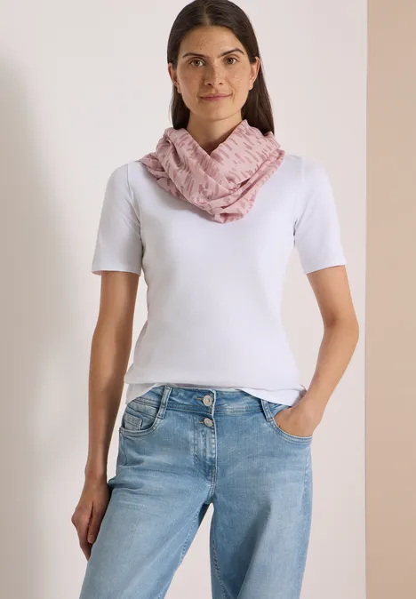 Une femme porte un t-shirt blanc, un jean et une écharpe rose, debout devant un fond clair.
