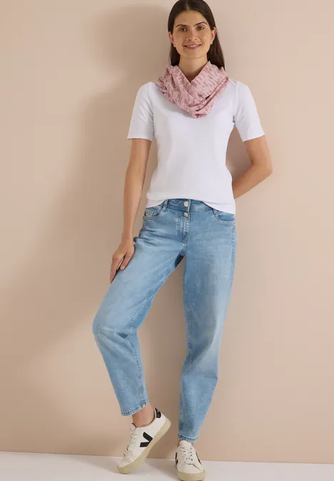 Femme en t-shirt blanc, jean bleu, foulard rose et baskets blanches, posant devant un mur neutre.