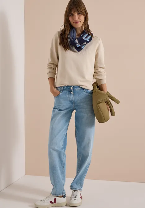 Femme en pull crème, écharpe bleue, jean ample et baskets blanches, tenant un sac vert.