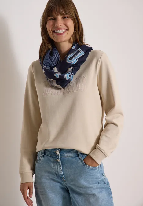 Femme souriante en pull beige, jean et foulard bleu.