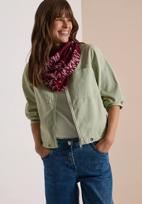 Femme en veste vert clair, jean et foulard rouge, souriante de profil.