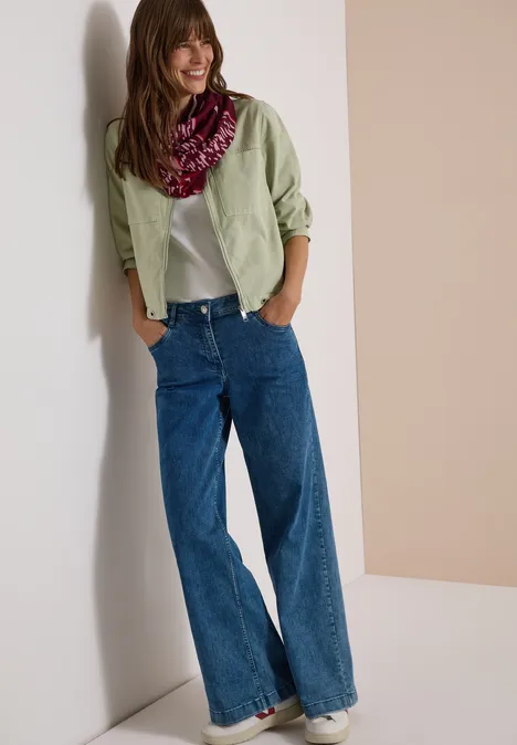 Femme souriante appuyée contre un mur, chemise vert pâle, t-shirt blanc, jean large bleu et foulard coloré.