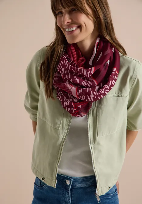 Femme en veste vert pâle, t-shirt blanc, jean et foulard bordeaux à motifs, souriante.
