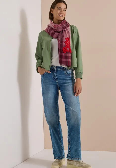 Vrouw in groene cardigan, paarse geruite sjaal en losse blauwe spijkerbroek tegen neutrale achtergrond.