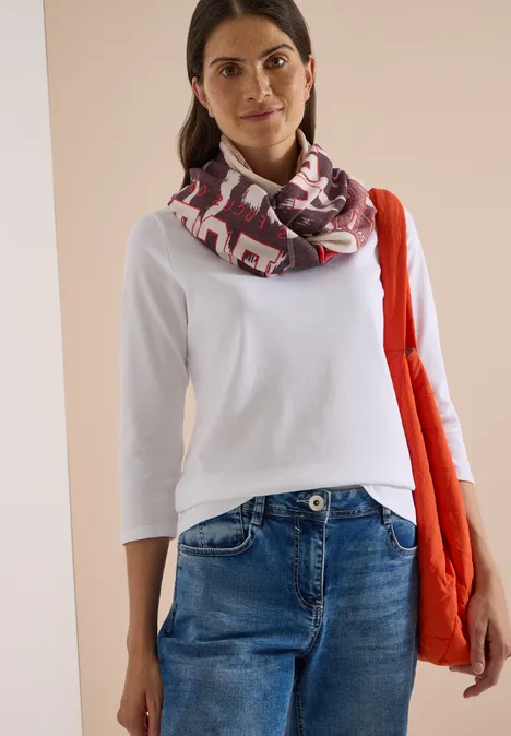 Femme portant un t-shirt blanc, un jean, une écharpe à motifs et un sac orange sur fond clair.