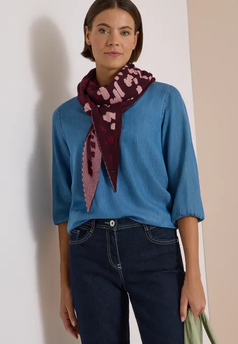 Frau in blauem Pullover, Jeans und lila Halstuch hält grüne Tasche.