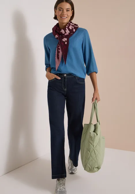 Frau in blauem Pullover, dunklen Jeans, Schal und Sneakers trägt eine grüne gesteppte Tasche.