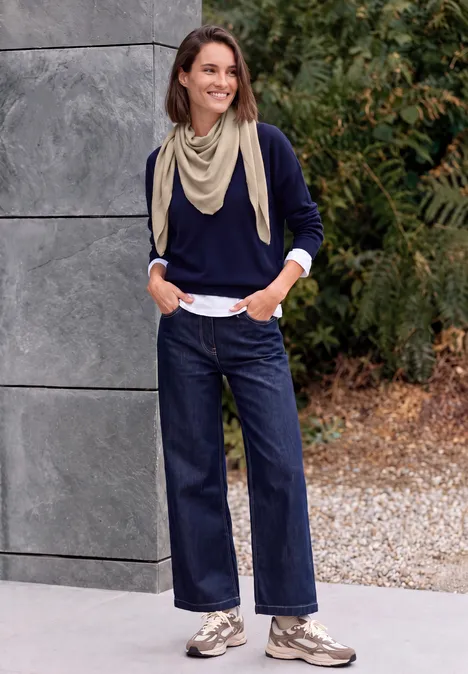 Eine Frau mit hellbeigem Schal, dunkelblauem Pullover, weiten Jeans und Sneakers vor einer Mauer mit Pflanzen.