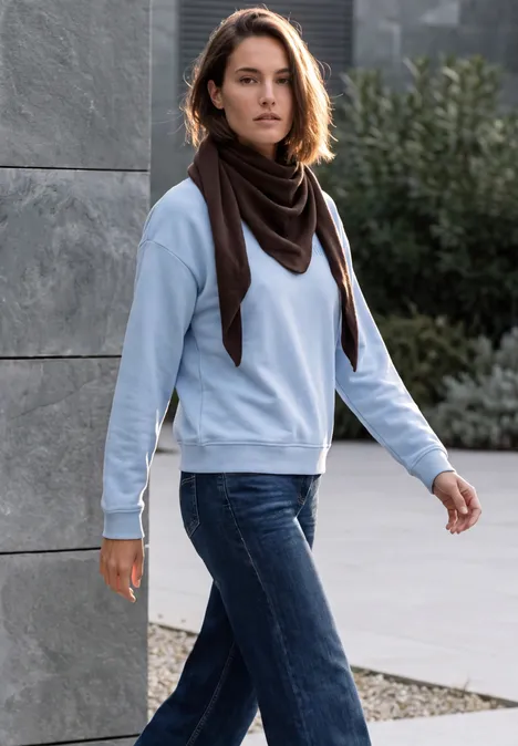 Dreieckstuch in Unifarbe truffle brown Frau in Jeans, hellblauem Pullover und dunklem Schal geht draußen an einer Betonwand vorbei.
