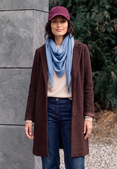 Eine Frau trägt einen braunen Mantel, blauen Schal, beigen Pullover, Jeans und eine burgunderfarbene Kappe.