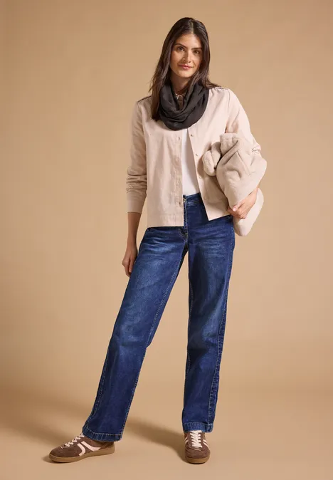 Frau in heller Strickjacke, schwarzem Loop, Jeans und Loafers vor neutralem Hintergrund.
