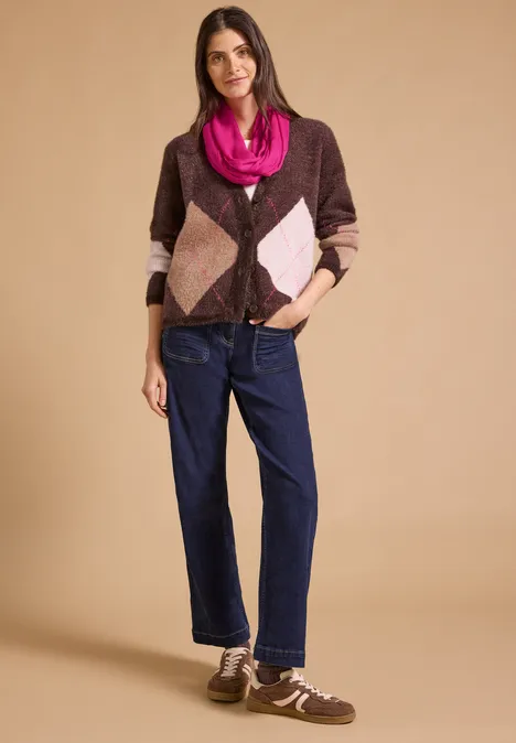 Frau trägt karierten Cardigan in Braun und Rosa, Jeans und Sneakers vor neutralem Hintergrund.