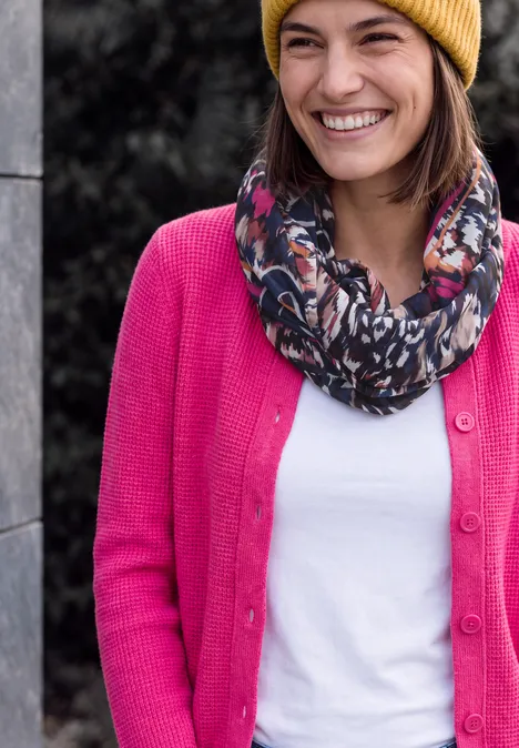 Loop-Schal mit Grafikprint truffle brown Eine Frau mit gelber Mütze, buntem Schal und pinkem Cardigan lächelt.