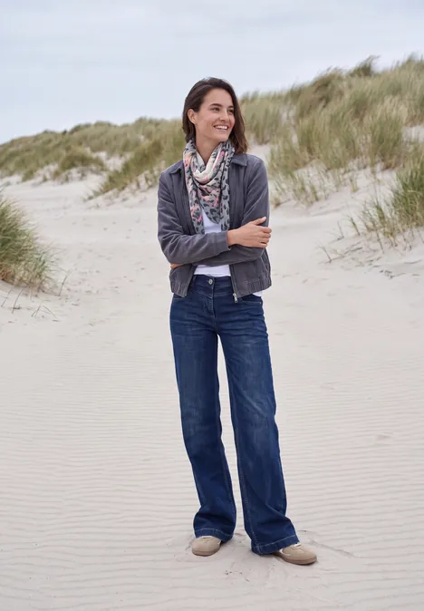 Une femme souriante en jean et foulard se tient détendue sur une plage de dunes herbeuses.
