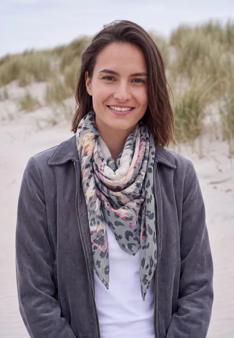 Femme souriante en écharpe et veste sur une plage de dunes, herbe verte en arrière-plan.